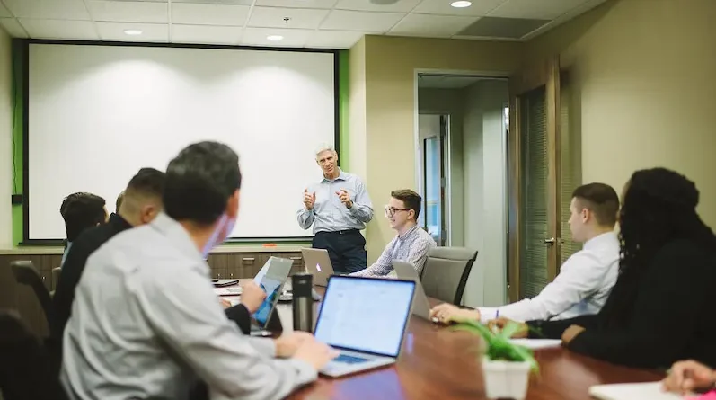 Business meeting in a modern conference room with attendees seated around a table, laptops open, and a presenter standing in front, displaying data on a screen.